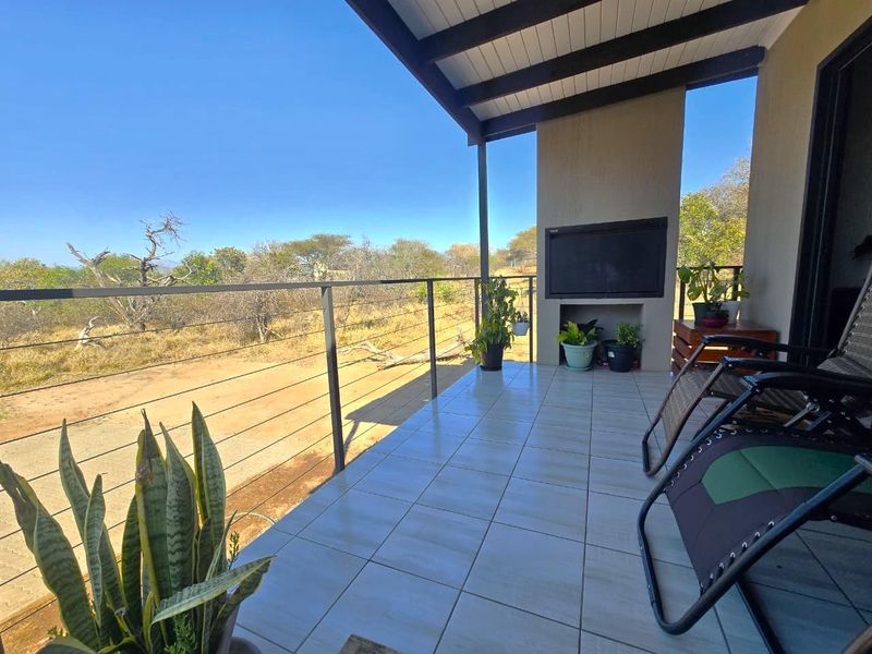 A covered outdoor patio featuring tiled flooring, a built-in braai, and a steel balustrade, offering expansive views of the surrounding natural landscape.