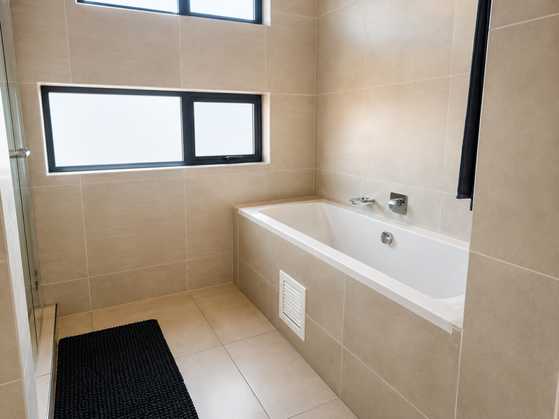 A modern, well-maintained bathroom featuring neutral floor-to-ceiling tiling, a built-in bathtub with wall-mounted fixtures, and a glass-enclosed shower area.