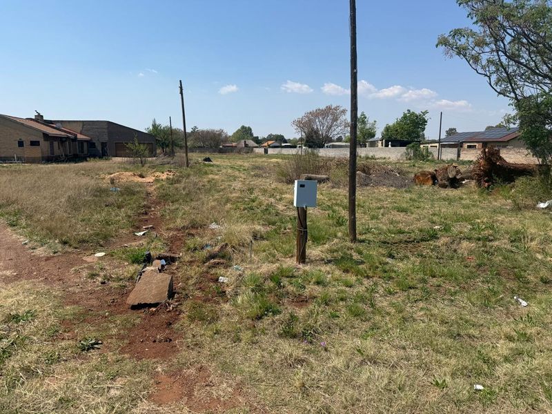 A vacant residential plot featuring an electrical utility box, utility pole, and surrounding boundary walls, with natural grass cover and some site debris.