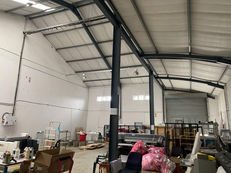 Spacious industrial warehouse unit featuring high ceilings with exposed steel trusses, concrete flooring, and a large roller shutter door for easy access.