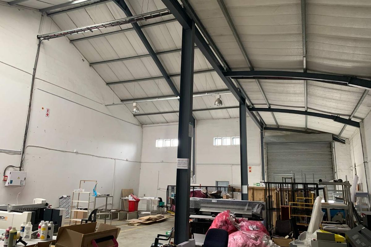 Spacious industrial warehouse unit featuring high ceilings with exposed steel trusses, concrete flooring, and a large roller shutter door for easy access.