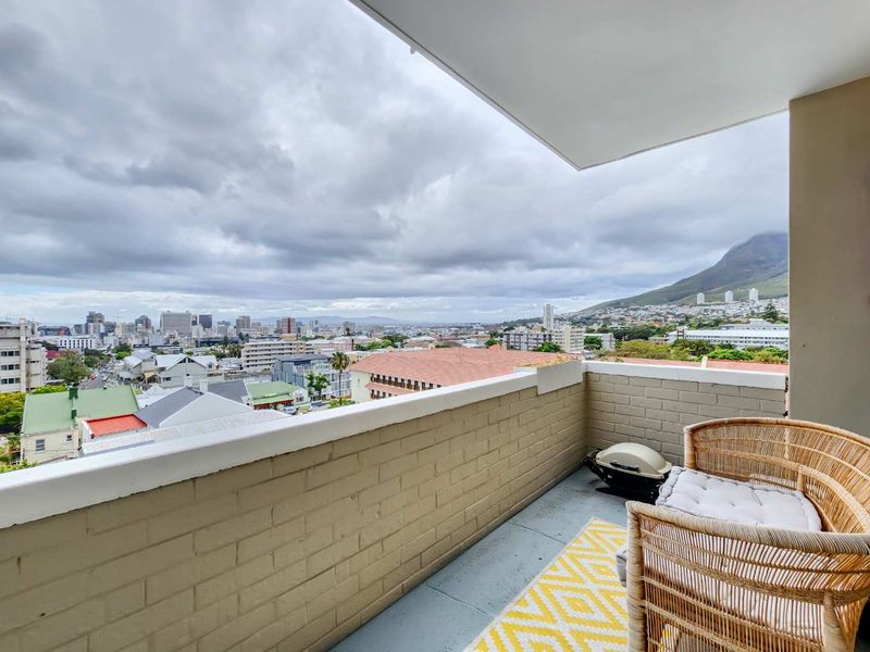A covered balcony featuring a painted brick parapet wall and concrete flooring, offering expansive views of the city and mountain landscape.