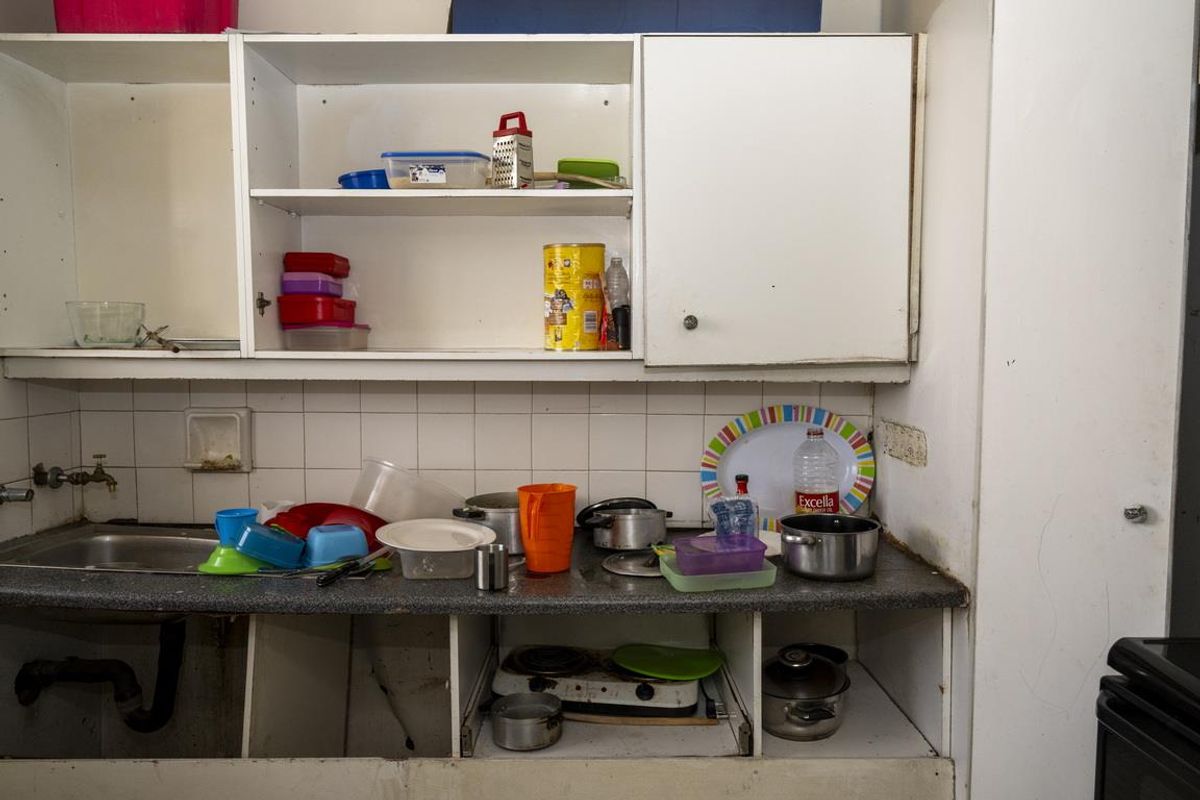 A dated kitchen area featuring basic cabinetry, a stainless steel sink, and tiled splashback, currently showing significant wear and tear.