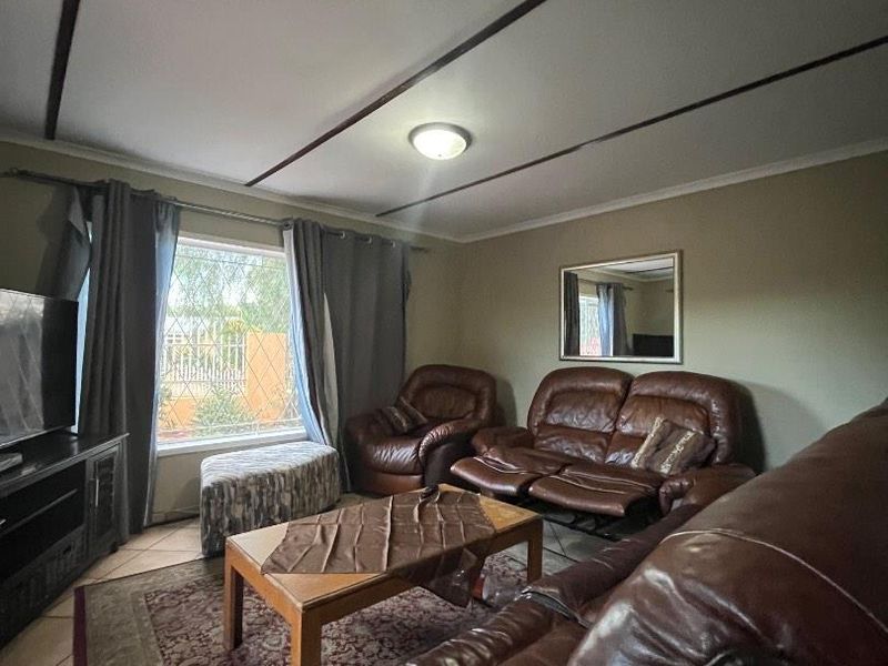 A comfortable living room featuring tiled flooring, ceiling beams, and a large window with security bars, providing a functional and well-lit space.