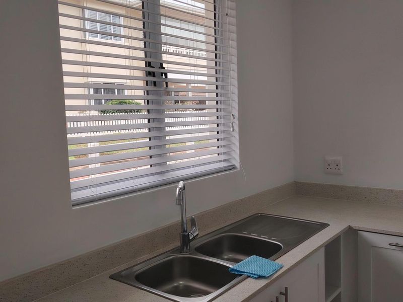 A clean, modern kitchen area featuring a double stainless steel sink, stone countertops, and white cabinetry with fitted venetian blinds.