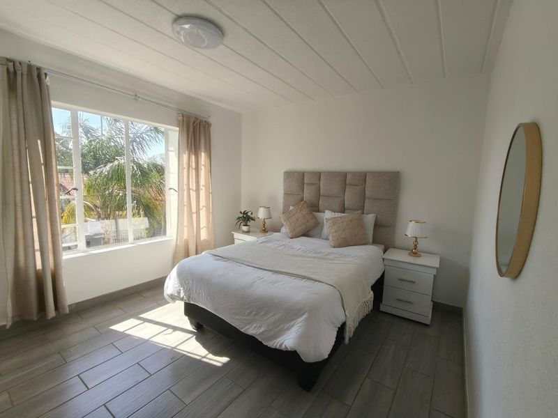 A bright and well-maintained bedroom featuring large windows, neutral tiled flooring, and a clean, modern aesthetic.