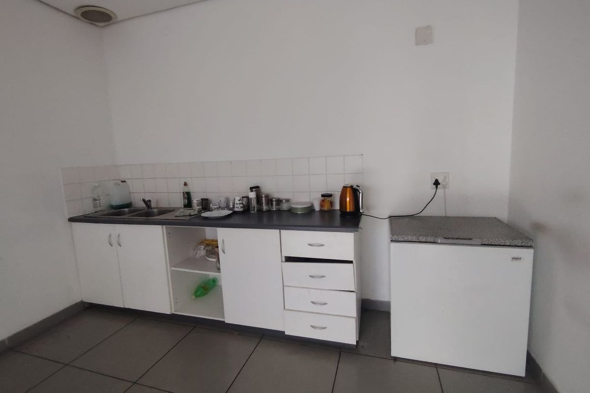 A compact kitchenette featuring white built-in cabinetry, a stainless steel sink, tiled splashback, and grey floor tiles in a functional retail space.