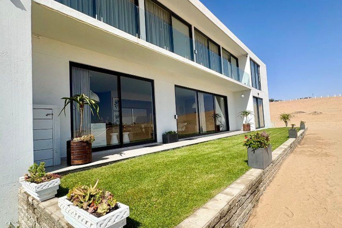 Rear exterior view of the townhouse featuring a manicured lawn, stone retaining wall, and a spacious balcony overlooking the sandy landscape.