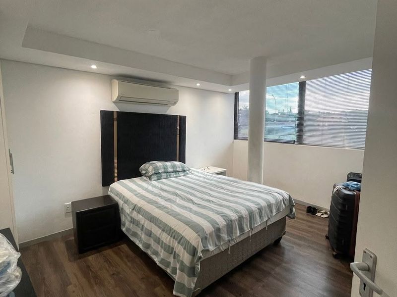 A well-lit bedroom featuring laminate flooring, a structural column, split-system air conditioning, and large windows with venetian blinds.