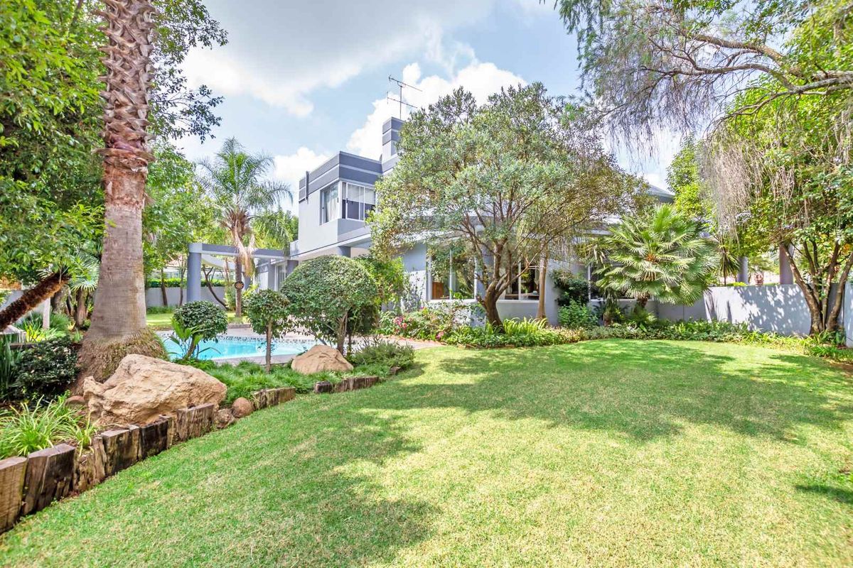A well-maintained rear garden featuring a lush lawn, mature trees, landscaped borders, and a private swimming pool adjacent to the house.