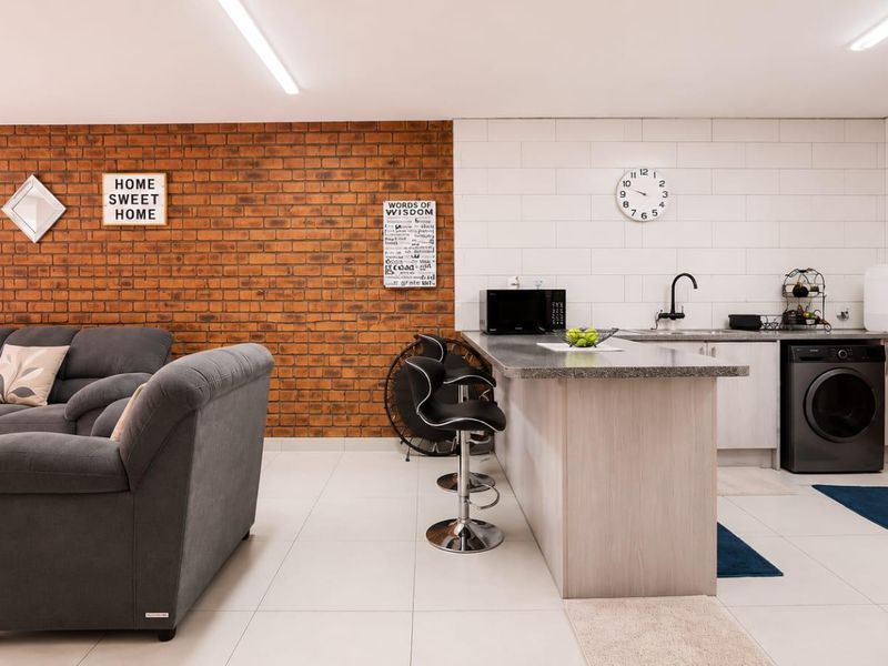 Open-plan living area featuring a kitchen island with sink, exposed brick accent wall, and modern tiled flooring throughout the combined space.