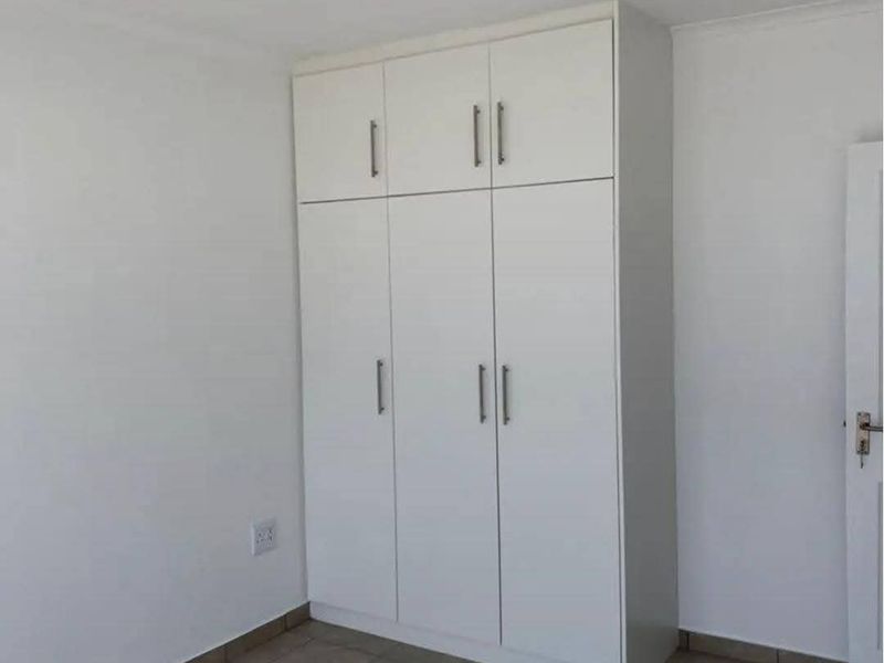 A clean, empty bedroom featuring neutral tiled flooring and a modern white built-in wardrobe with overhead storage compartments.