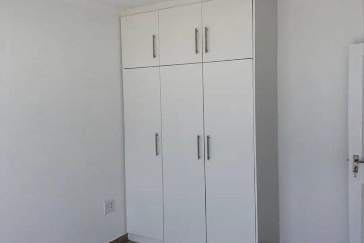 A clean, empty bedroom featuring neutral tiled flooring and a modern white built-in wardrobe with overhead storage compartments.