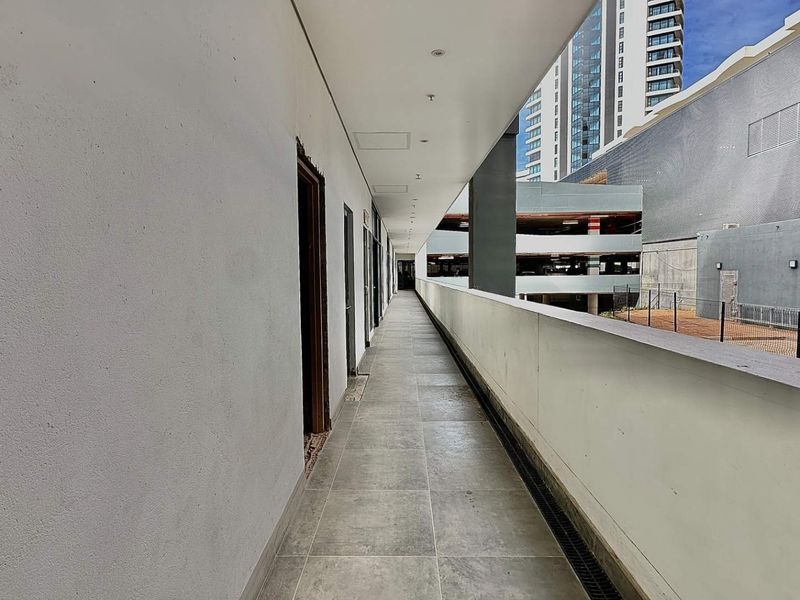 A covered exterior walkway or balcony featuring tiled flooring, recessed ceiling lighting, and a protective balustrade overlooking the surrounding urban office complex.