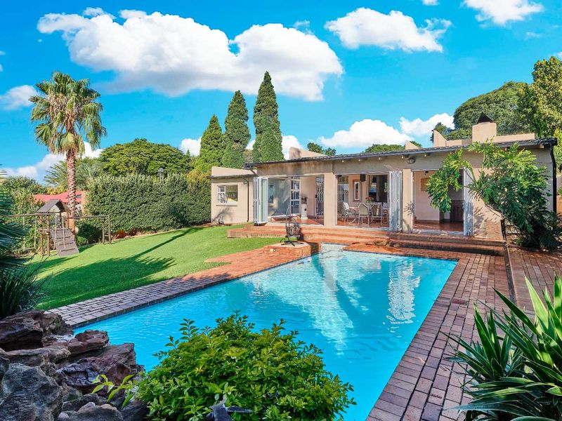Rear view of the property featuring a swimming pool, brick-paved patio area, and a well-maintained garden with folding glass doors leading into the home.
