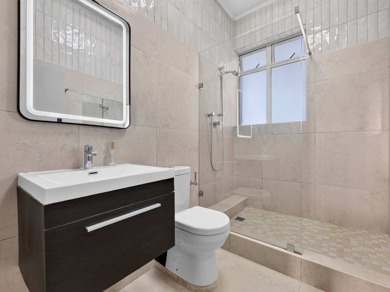 Modern bathroom featuring a dark wood vanity, backlit mirror, walk-in shower with glass screen, and neutral floor-to-ceiling tiling.