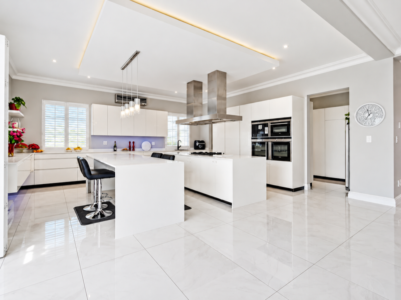 Modern, high-end kitchen featuring a large central island, integrated stainless steel appliances, sleek white cabinetry, and polished porcelain flooring with recessed ceiling lighting.