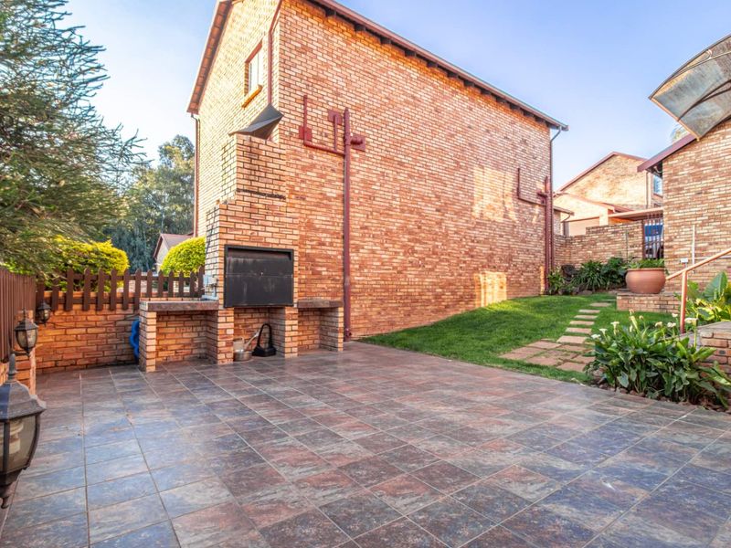 A private rear courtyard featuring a paved patio area with a built-in brick braai, lawn, and established garden borders.