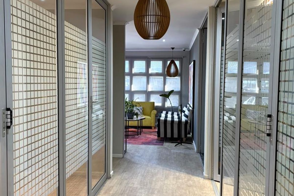 A modern office hallway featuring glass partition walls with frosted privacy detailing and wood-look laminate flooring, leading into an open-plan workspace.