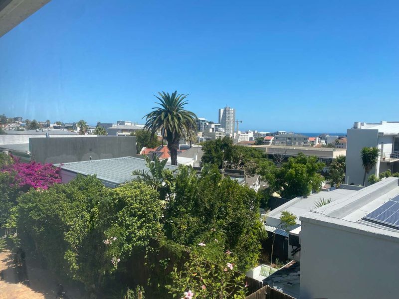 An elevated urban view overlooking surrounding residential rooftops, lush greenery, and distant city buildings under a clear blue sky.