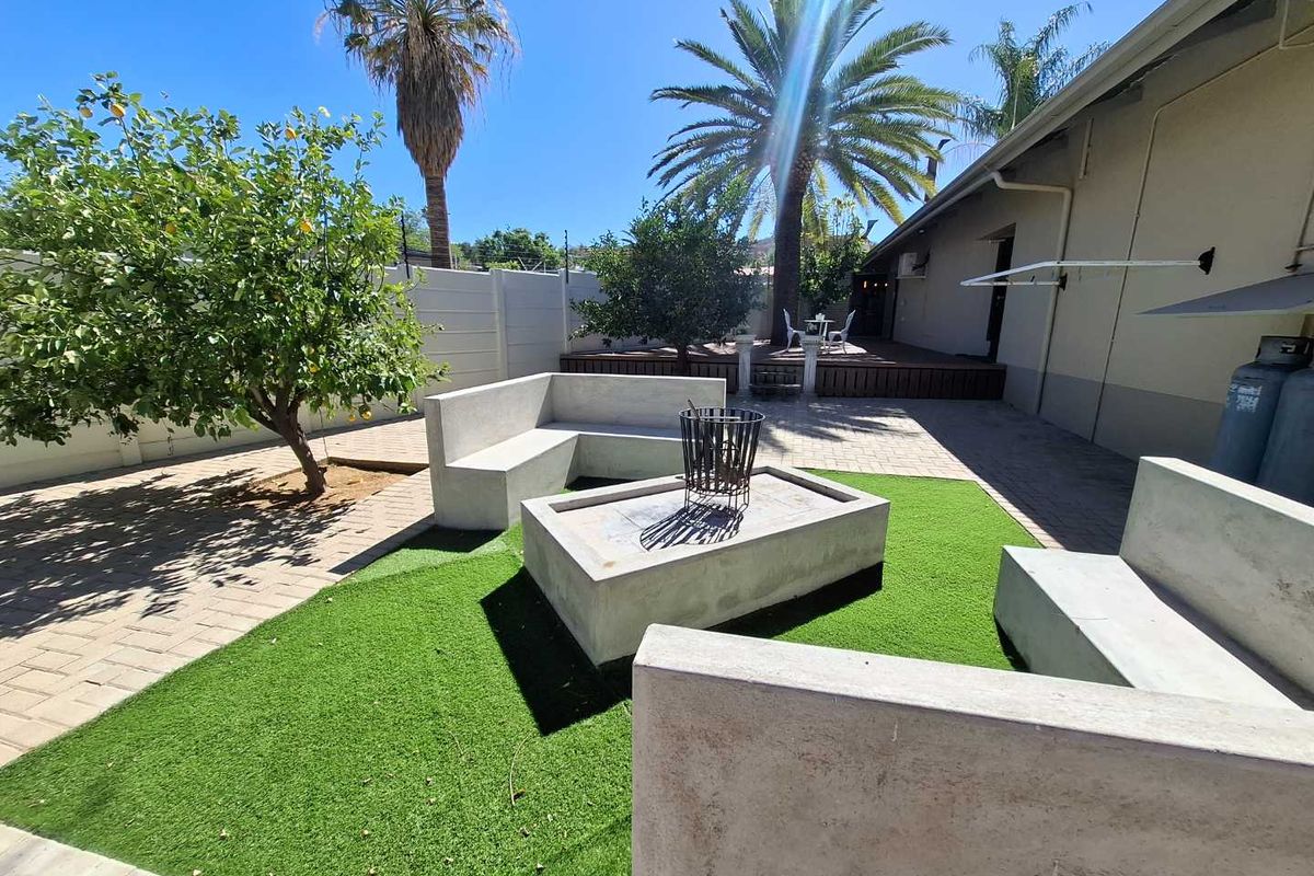 A well-maintained rear courtyard featuring a paved patio, artificial lawn, built-in concrete seating with a fire pit, and a raised timber deck area.