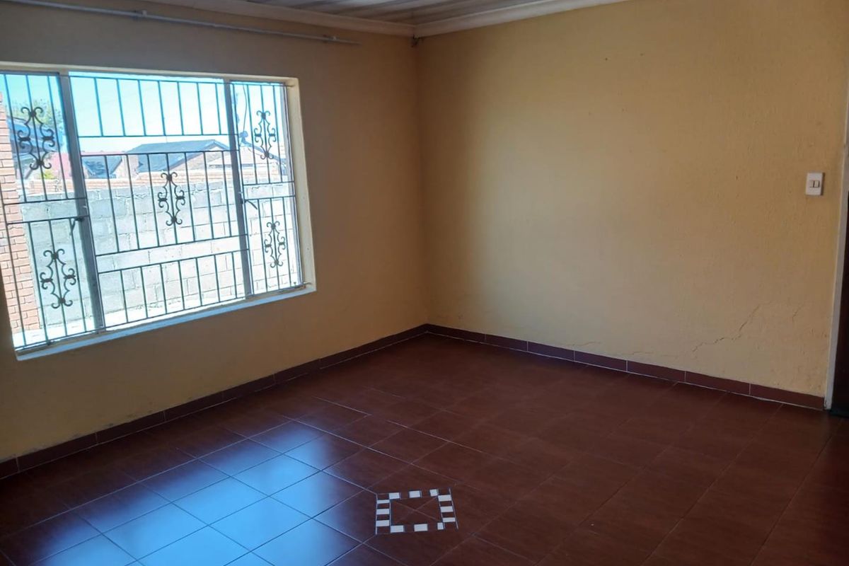 An empty living room featuring tiled flooring, security bars on the window, and a corrugated ceiling, requiring minor cosmetic updates.