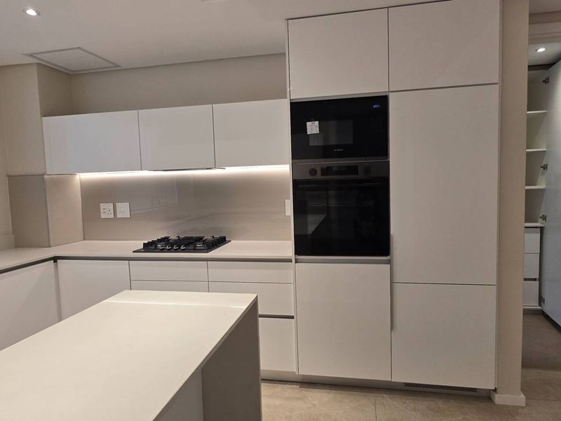 Modern, minimalist kitchen featuring handleless white cabinetry, stone countertops, integrated appliances, and a gas hob, with an adjacent walk-in pantry.