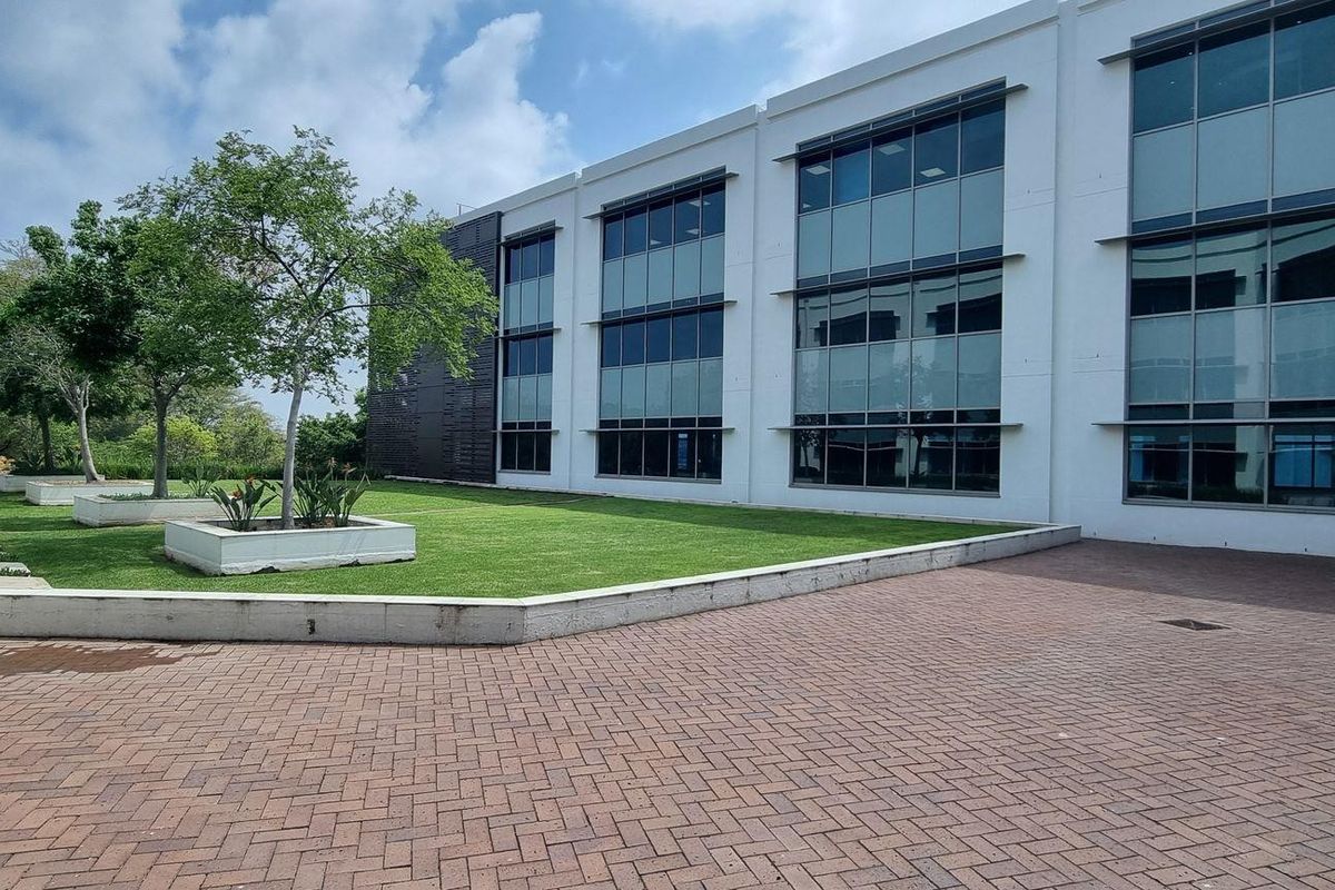 Modern office building exterior featuring a paved courtyard, landscaped garden beds, and large glass curtain wall windows.