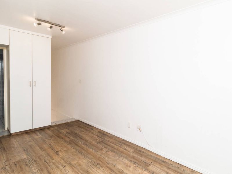 An empty bedroom featuring laminate flooring, built-in cupboards, and track lighting, with an adjacent view into a tiled bathroom.