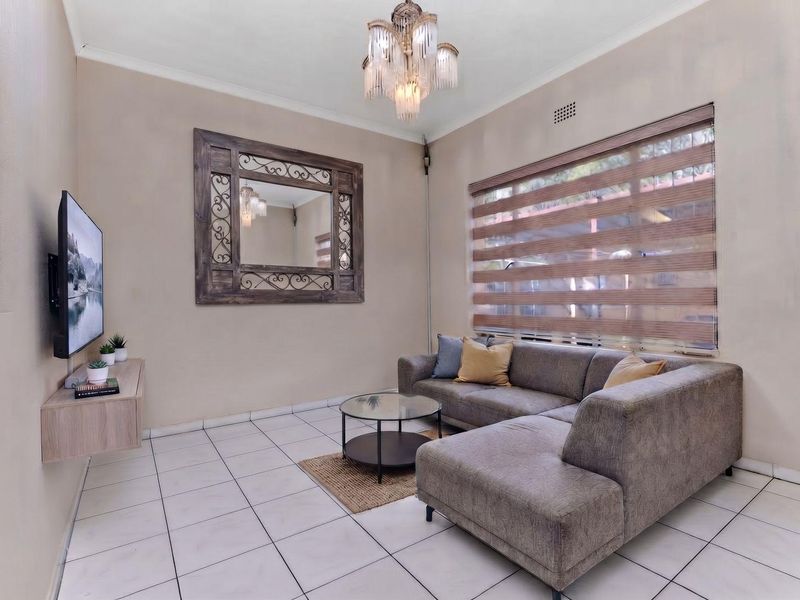 A well-maintained living room featuring tiled flooring, a decorative chandelier, and a large window with modern fitted blinds.