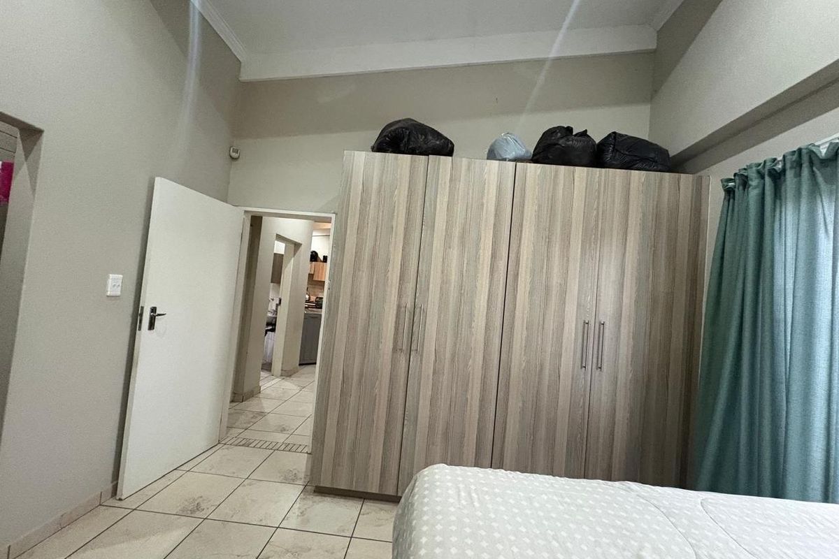 A bedroom featuring built-in wooden cupboards, tiled flooring, and high ceilings, with an adjacent view into the hallway and kitchen area.