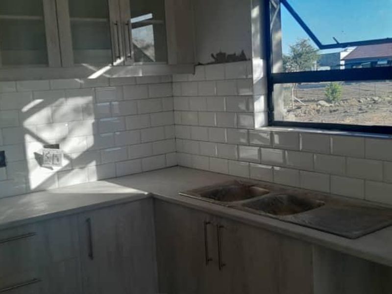 An unfinished kitchen area featuring light wood-look cabinetry, a double basin sink, and a white subway tile backsplash, awaiting final wall finishing and electrical installation.