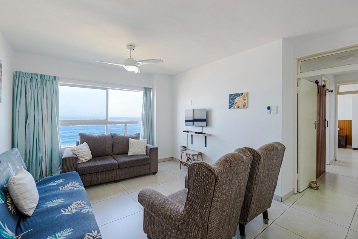 A bright living room featuring large windows with ocean views, tiled flooring, and a ceiling fan, providing a comfortable and airy living space.