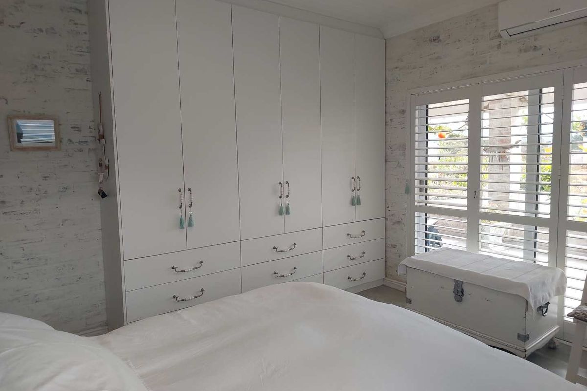 A bright bedroom featuring a large built-in wardrobe, split-system air conditioning, and plantation shutters, complemented by a textured exposed brick wall.