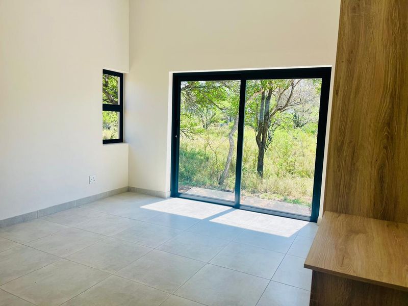 A bright, modern bedroom featuring neutral tiled flooring, built-in cabinetry, and large sliding glass doors that provide natural light and views of the surrounding greenery.