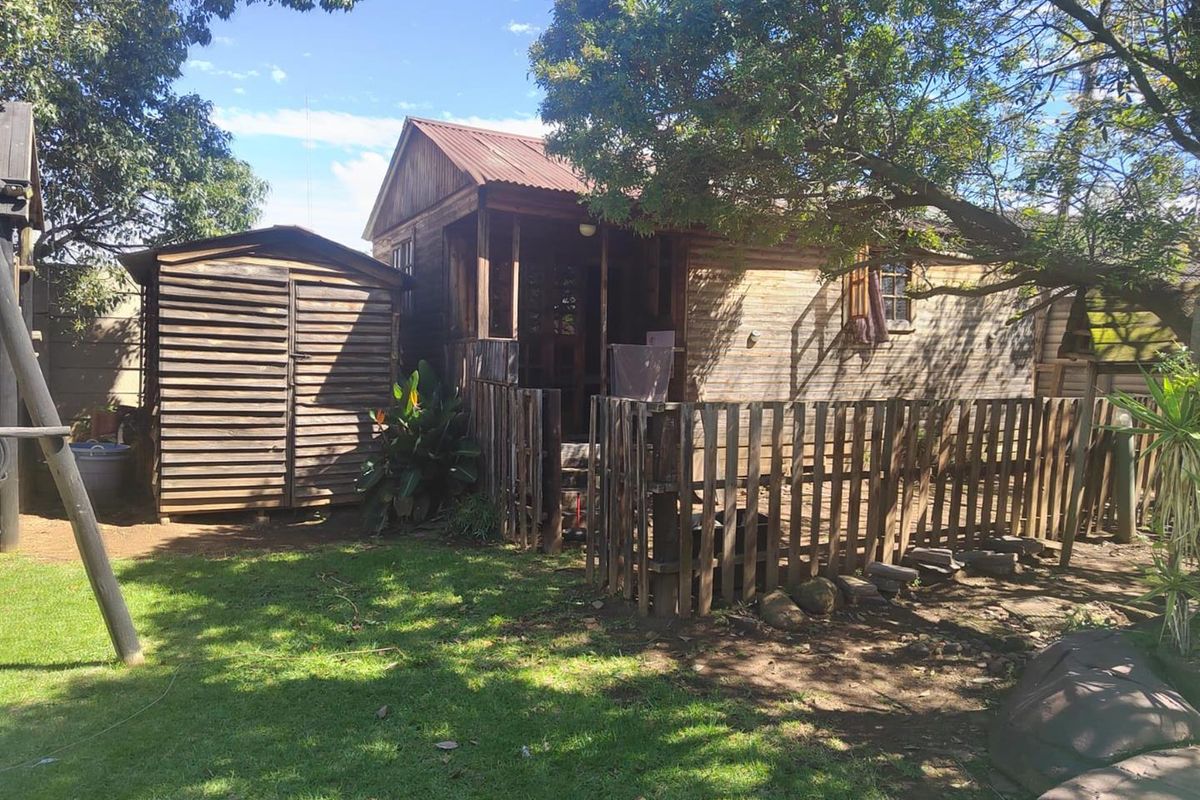 A rustic timber-clad dwelling featuring a small attached deck, corrugated metal roof, and an adjacent wooden storage shed set within a garden area.