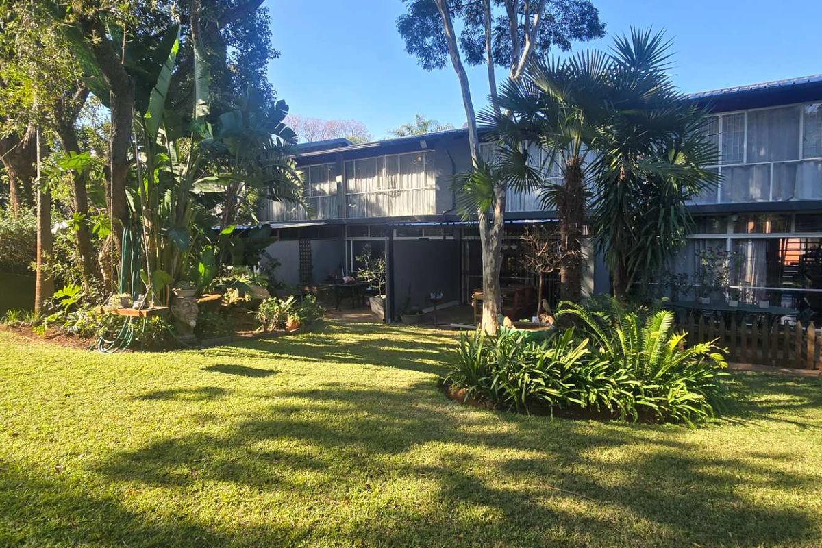 A view of the rear elevation of the townhouse, featuring a spacious lawn, mature garden landscaping, and a covered patio area.