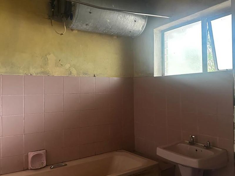 A dated bathroom requiring significant renovation, featuring a pink bathtub and pedestal sink, with visible wall damage and exposed water heating infrastructure.