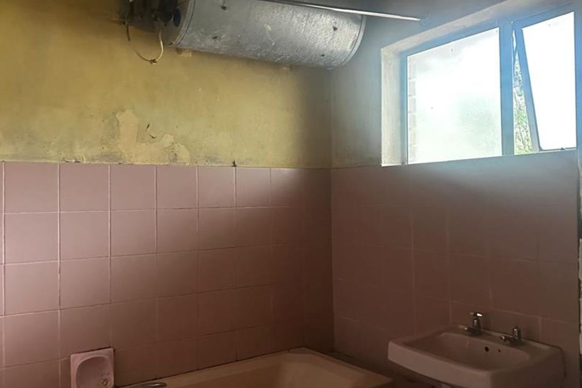 A dated bathroom requiring significant renovation, featuring a pink bathtub and pedestal sink, with visible wall damage and exposed water heating infrastructure.