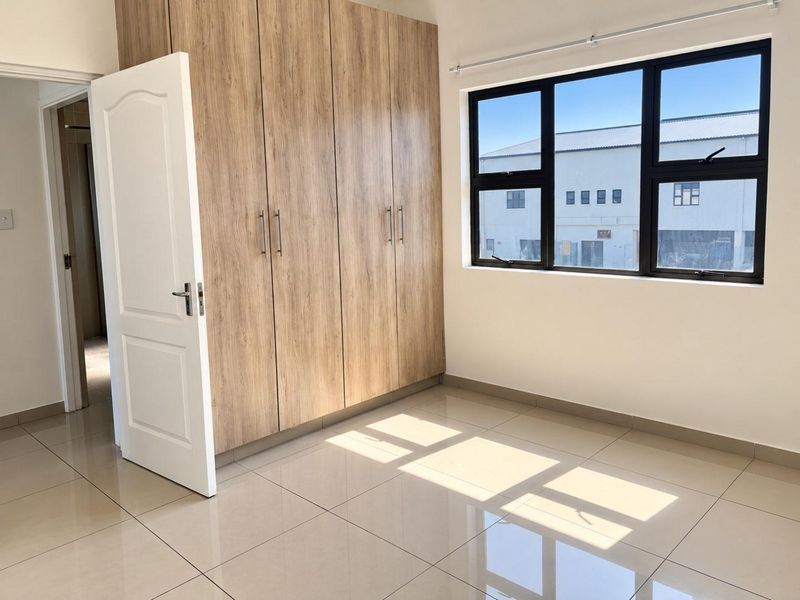 A bright, empty bedroom featuring modern wood-finish built-in cupboards, neutral tiled flooring, and large aluminium-framed windows.