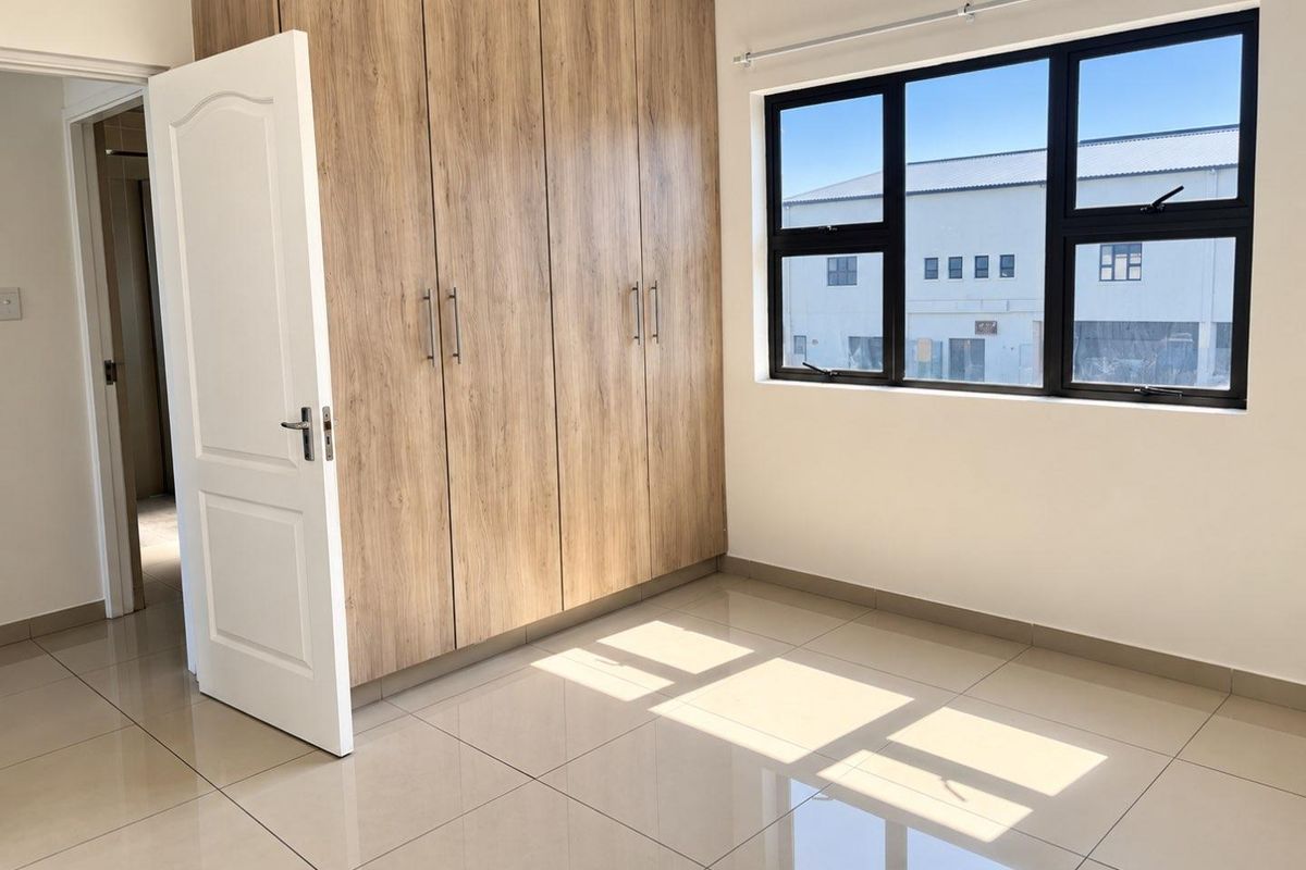A bright, empty bedroom featuring modern wood-finish built-in cupboards, neutral tiled flooring, and large aluminium-framed windows.