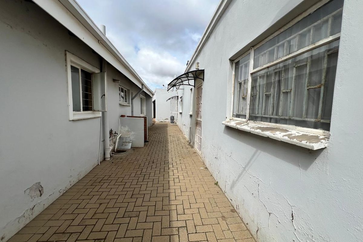 A narrow paved side passage between two building sections featuring security bars on windows and exterior wall maintenance required.