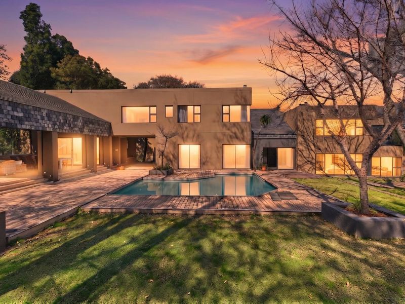 Rear view of a modern multi-level house featuring a swimming pool, timber decking, and expansive windows set within a landscaped garden at sunset.