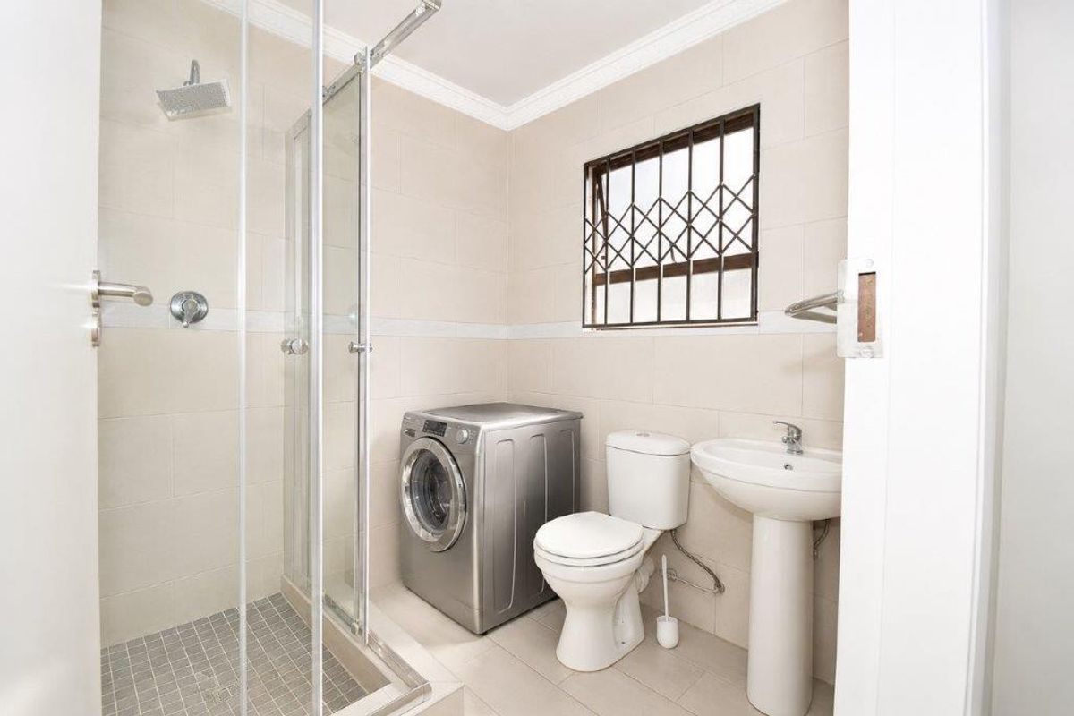 A functional bathroom featuring a glass-enclosed shower, pedestal basin, toilet, and integrated laundry space with tiled walls and flooring.