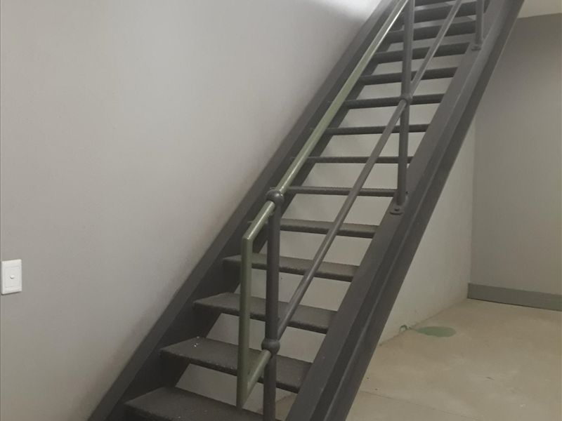 A functional industrial-style metal staircase with a matching handrail, set within a minimalist space featuring concrete flooring and neutral grey walls.