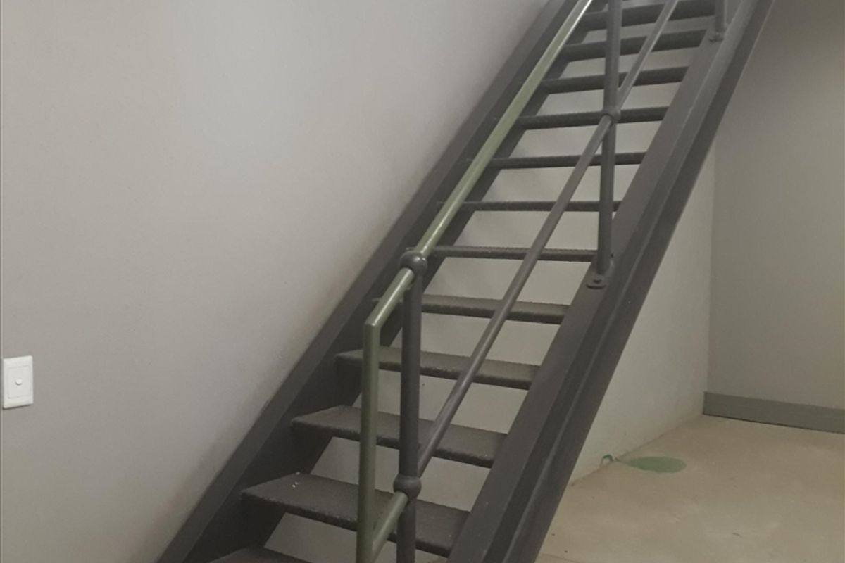 A functional industrial-style metal staircase with a matching handrail, set within a minimalist space featuring concrete flooring and neutral grey walls.