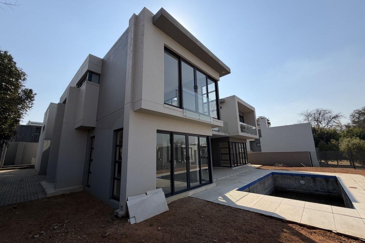 Modern double-storey rear elevation featuring large glass sliding doors, balcony with glass balustrades, and an adjacent swimming pool set in an unfinished garden.