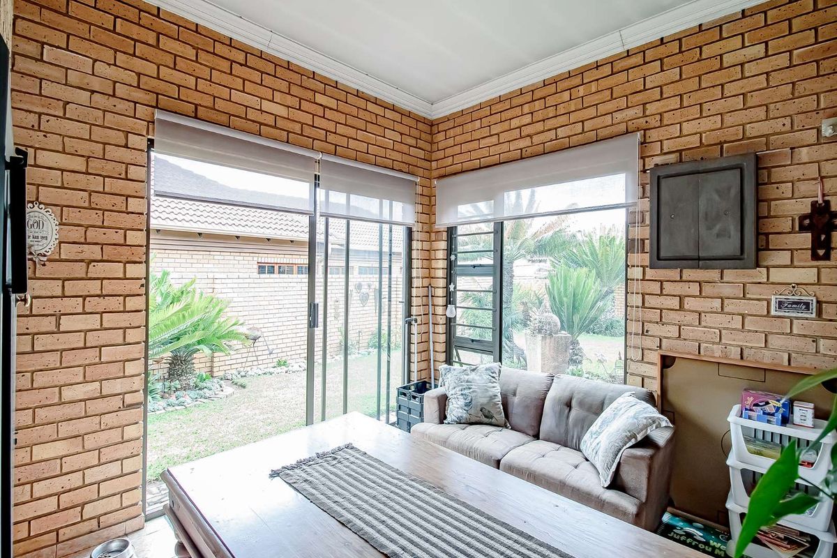 A sunroom or living area featuring exposed face-brick walls, large sliding glass doors, and roller blinds, offering a view of the rear garden.