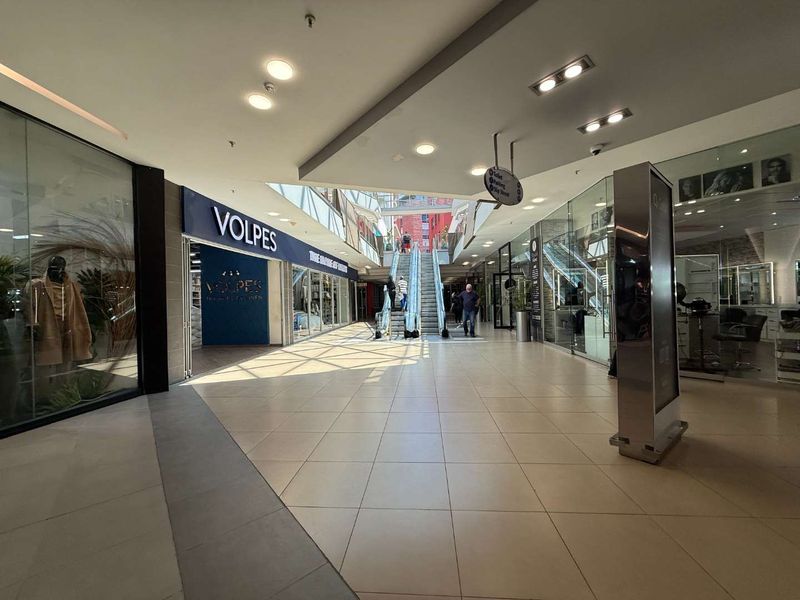 Interior view of a modern retail shopping center corridor featuring tiled floors, glass storefronts, and an escalator leading to the upper level.