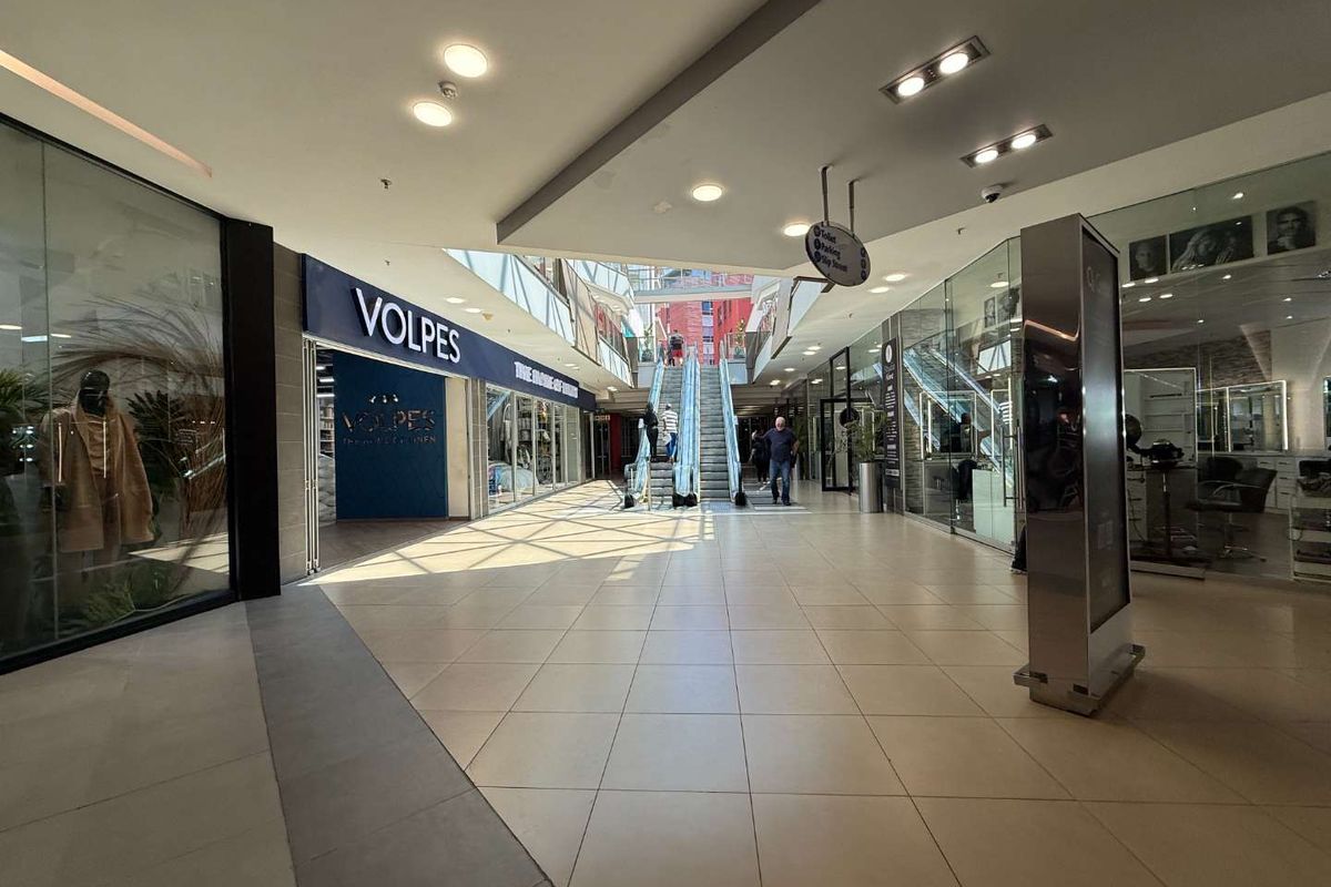 Interior view of a modern retail shopping center corridor featuring tiled floors, glass storefronts, and an escalator leading to the upper level.